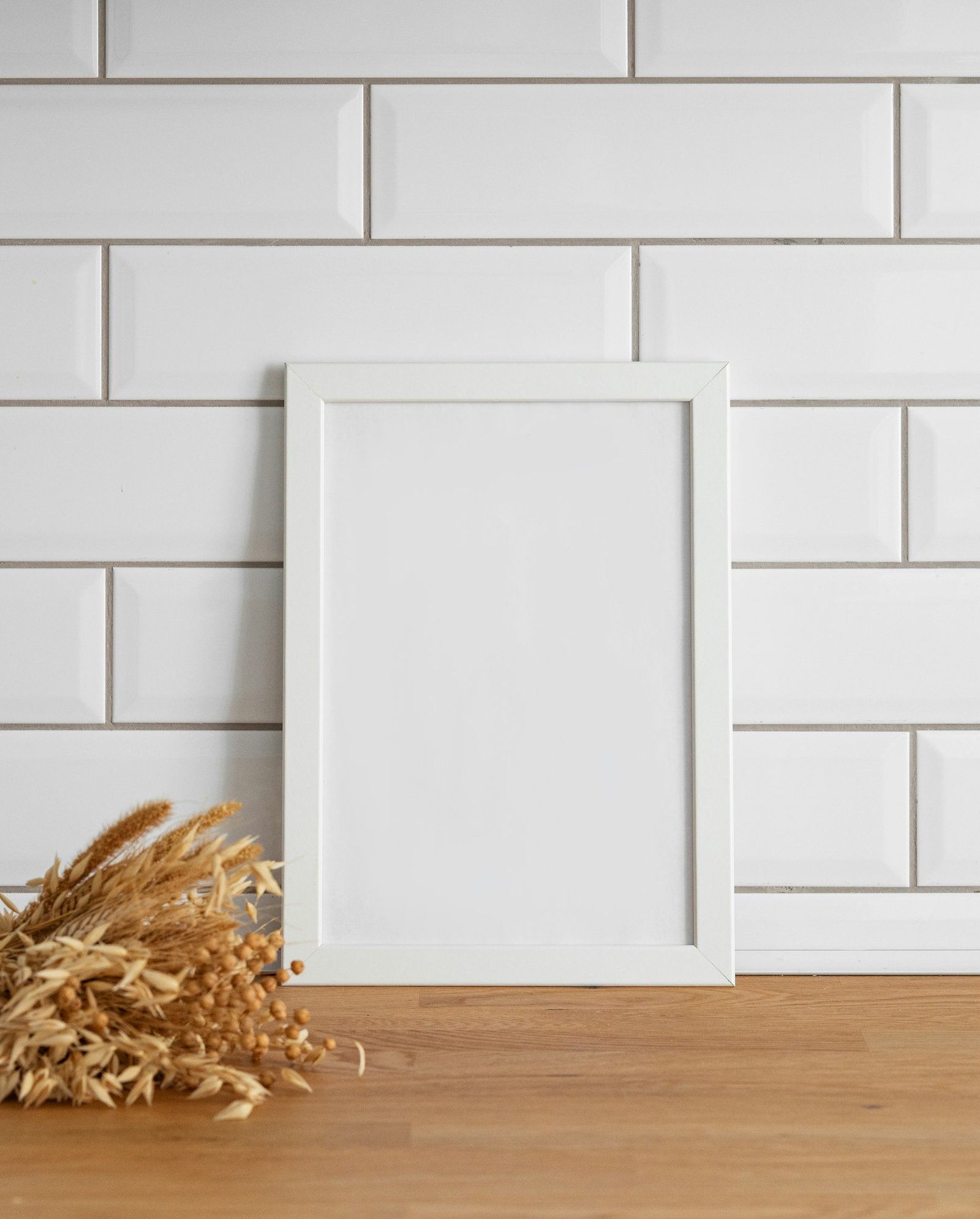 A frame with a blank canvas against a white tile wall and on a wooden tabletop with dry bouquet.